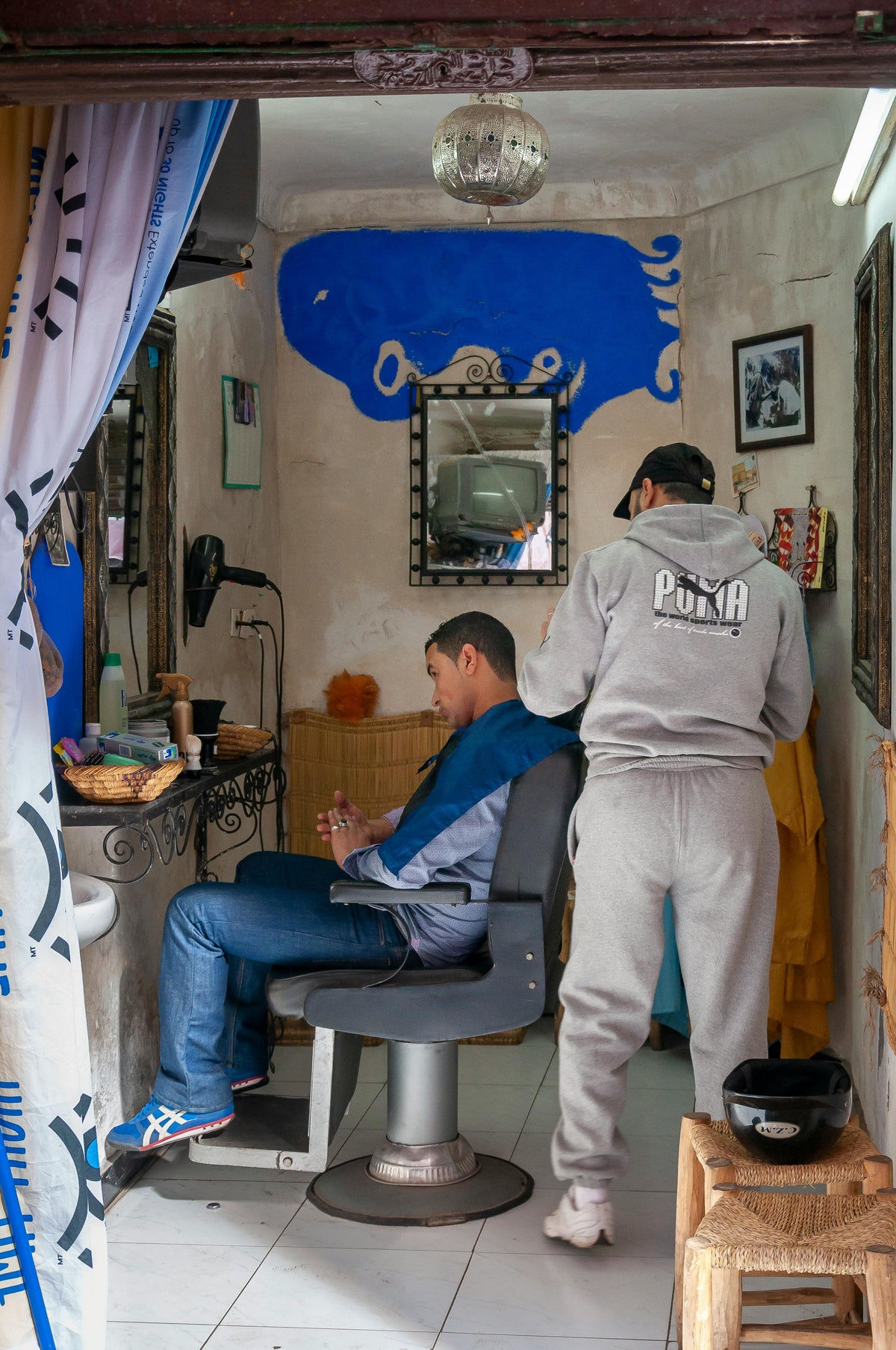Anonymous man in casual clothes cutting hair of young ethnic male client in rustic traditional barbershop